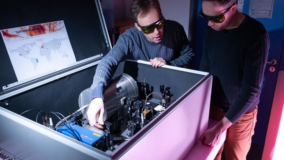 GNOME-Messstation am Leibniz-IPHT in Jena: Dr. Theo Scholtes (links) und Göran Hellmann justieren Quantenmagnetometer zur Suche nach Spuren Dunkler Materie. Im Dauerbetrieb sendet er seine Messdaten an den GNOME-Server. Messtation: Kasten in den Kabel hereinlaufen, technische Komponenten miteinander verknüpft sind und zwei Männer mit schwarzen Brillen, die sich unzerhalten und einen Mechanismus bedienenn