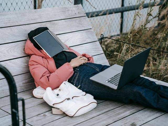 Frau in rosa Jacke liegt schlafend mit Laptop auf dem Schoß im Freien.