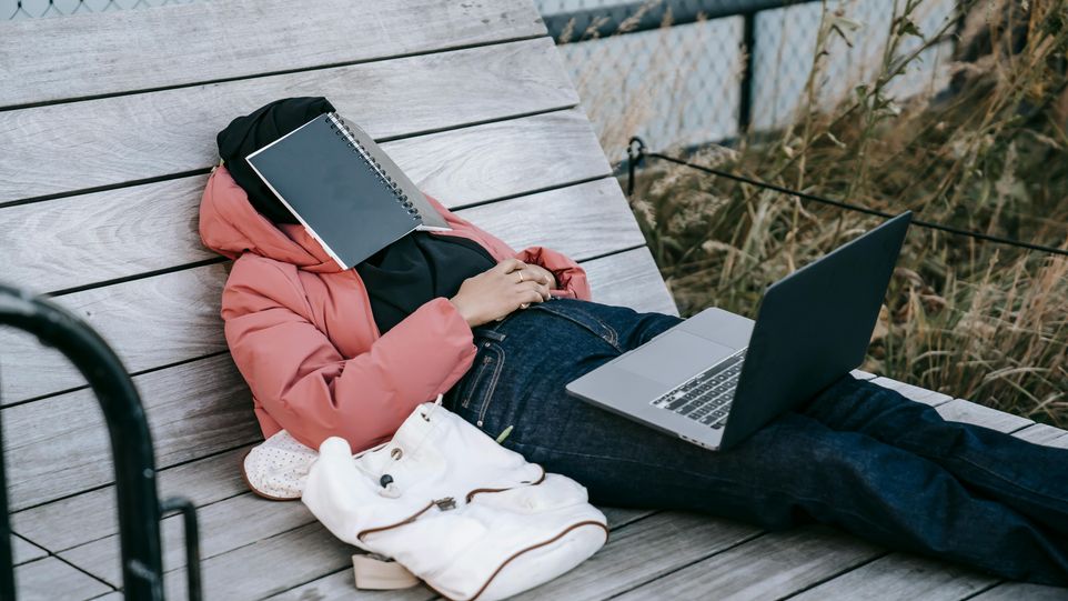 Frau in rosa Jacke liegt schlafend mit Laptop auf dem Schoß im Freien.