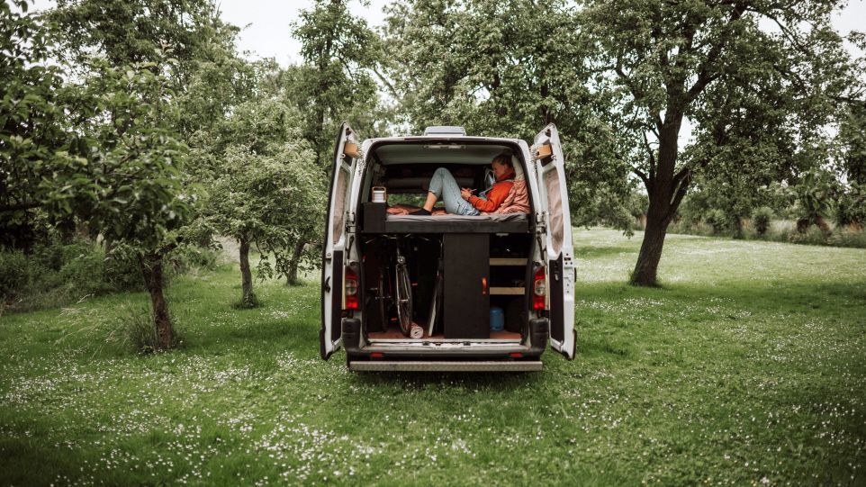 Zum Wohnwagen umgebauter Kleinbus in grüner Tür. Durch die geöffnete Tür sieht man eine lesende junge Frau. 