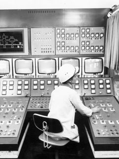 In einem Kontrollraum sitzt eine Person vor einem großen Bedienpult mit zahlreichen Schaltern, Anzeigen und Bildschirmen. Die Schwarzweißaufnahme zeigt einen technischen Schaltraum in einem Industriebetrieb.