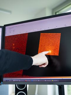 Eine Hand zeigt auf einen leuchtenden Punkt in einer farbigen rot-orangenen Mess- oder Bilddarstellung auf einem Computerbildschirm, flankiert von weiteren Monitoren. 