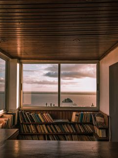 Innenraum mit Holzdecke und Bücherregalen vor einem großen Fenster, das auf Meer, Himmel und ein Schiff blickt.