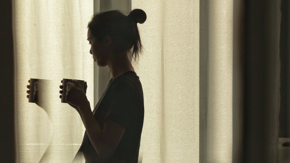 Frau vor mit Tasse vor verhangenem Fenster