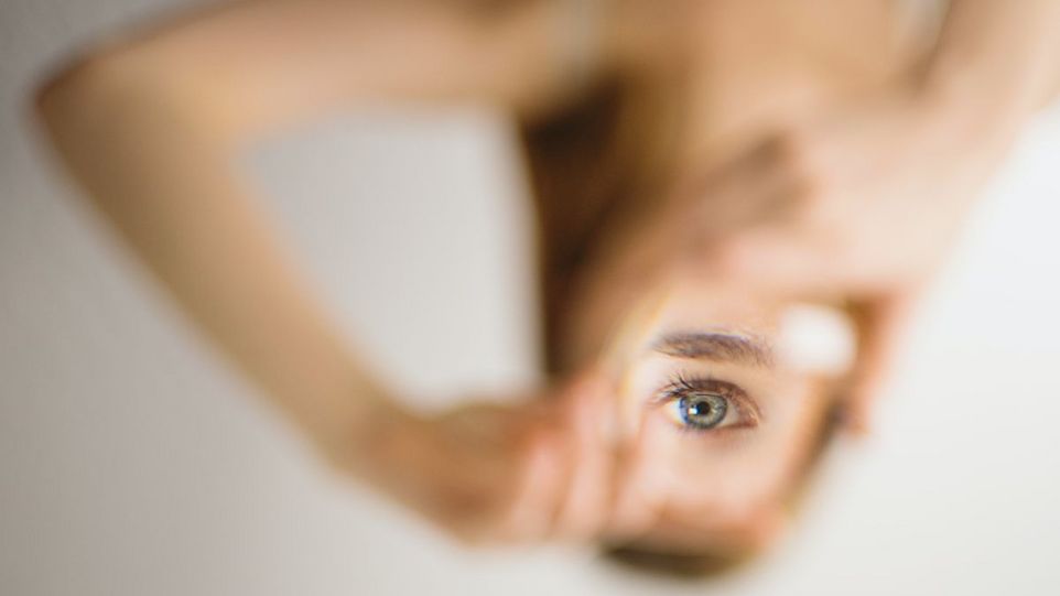 Hände, die einen Spiegel halten, in dem sich das Auge einer Frau spiegelt.