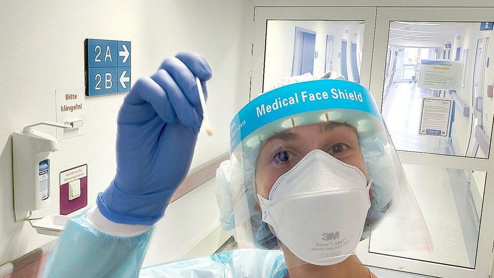Weibliche Pflegekraft mit Medical Face Shield und Maske, die ein Teststäbchen in die Kamera hält.