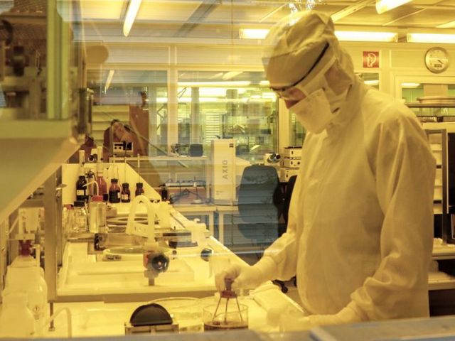 Person mit Sicherheitsanzug und Brille steht im Labor und führt Experiment durch. 