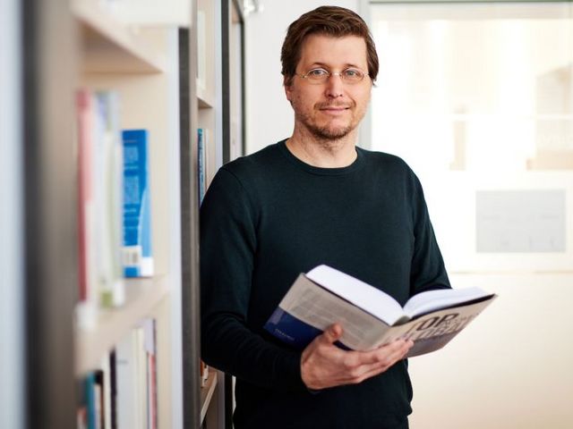 Moritz Schularick vor einem Bücherregal, ein aufgeschlagenes Buch in der Hand.