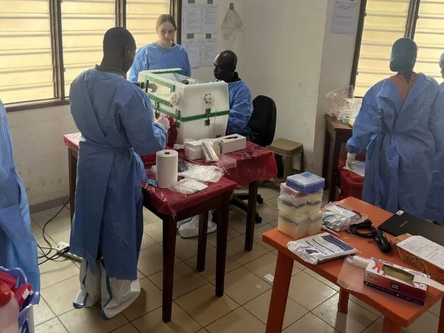 Foto von medizinischen Fachkräften in blauer Schutzkleidung, die in einem Laborraum an verschiedenen medizinischen Geräten arbeiten