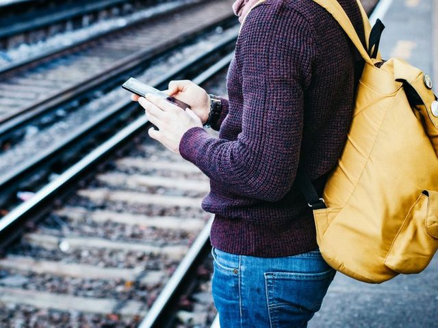 Junger Mann mit Smartphone, an einem Bahnhofsgleis stehend