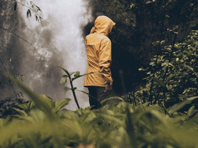 Rückenansicht einer Person im Regenmantel, vor einem im Wald gelegenen Wasserfall