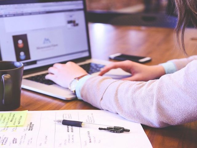 Frau arbeitet an Laptop, Kaffeetasse, Smartphone und Notizblock liegen daneben