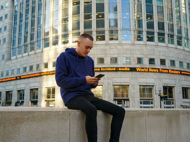Junger Mann sitzt auf einer Mauer vor einem Banner mit Reuters Nachrichten und schaut auf sein Smartphone.