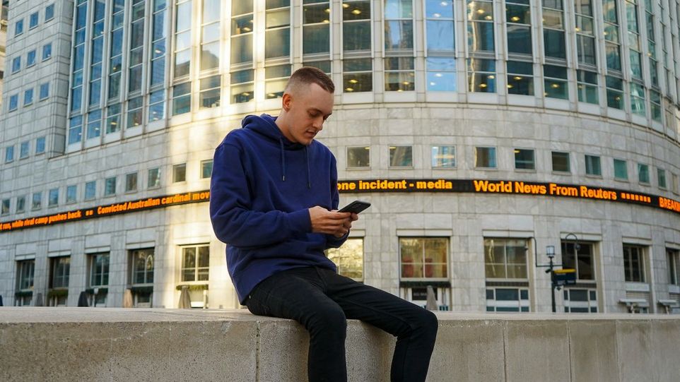 Junger Mann sitzt auf einer Mauer vor einem Banner mit Reuters Nachrichten und schaut auf sein Smartphone.