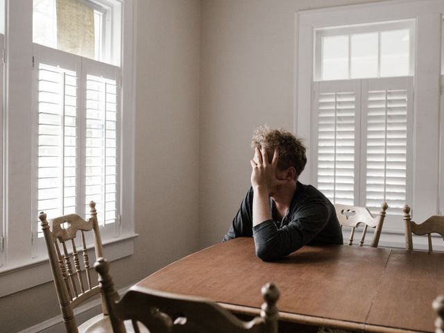 A man is sitting alone in a room at a table, his face in his hand.