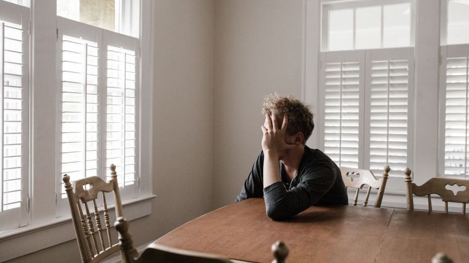 A man is sitting alone in a room at a table, his face in his hand.