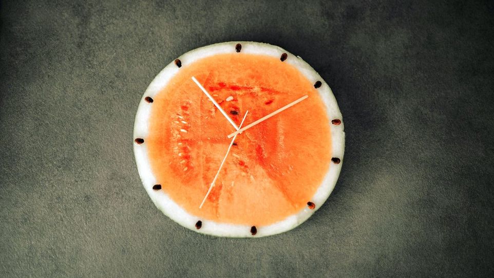 Beim Intervallfasten wird in einem bestimmten Rhythmus zwischen Zeiten der Nahrungsaufnahme und des Fastens gewechselt. Aufgeschnittene Melone, mit Holzzeigern als Uhr drapiert.