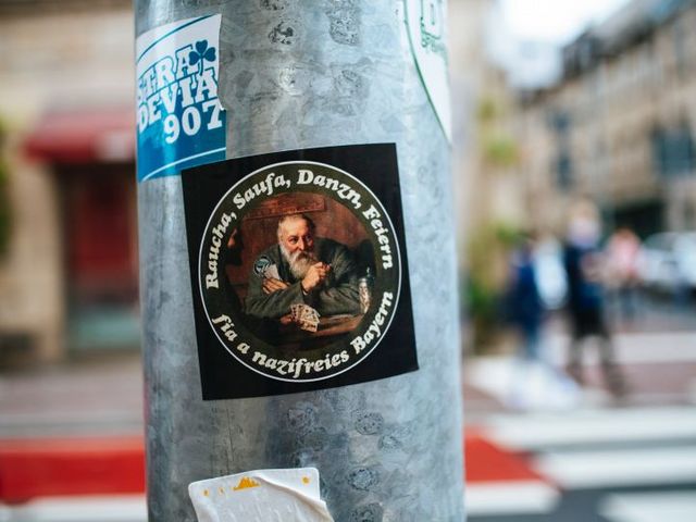 Aufkleber auf Straßenpfahl mit der Aufschrift: Raucha, Saufa, Danzn, Feiern - fia a nazifreies Bayern.